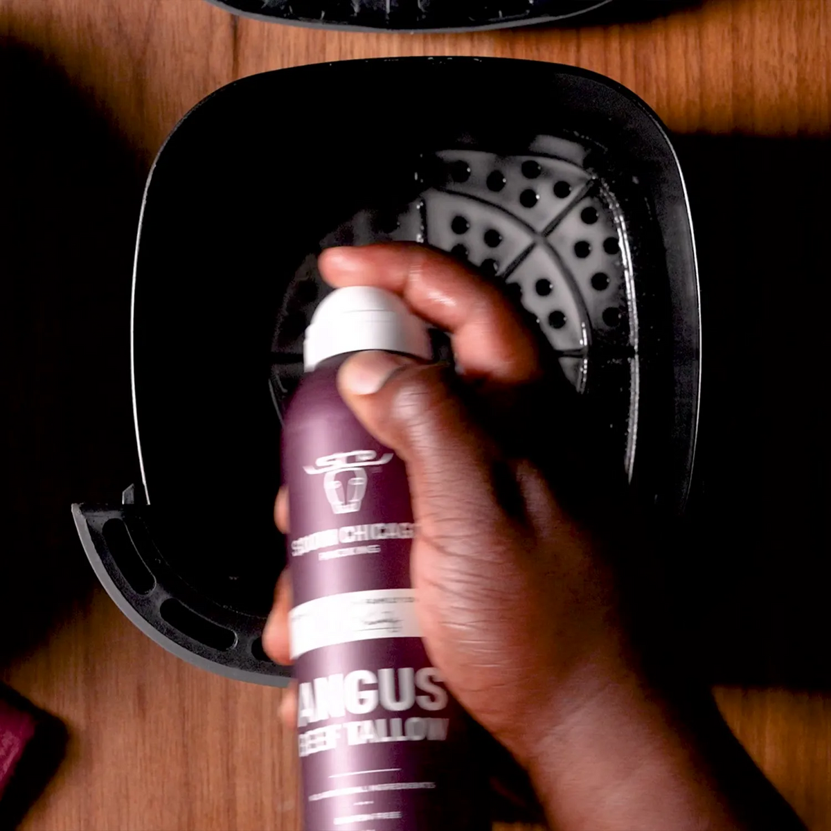 A hand holding a bottle of Chicago Cleaning Company Angus Beef Tallow spray, spraying inside a black air fryer basket on a wooden table.