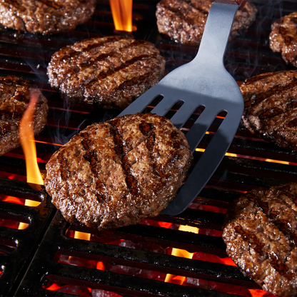 A black metal burger spatula lifting a grilled beef patty on a grill with other patties, flames, and smoke visible.