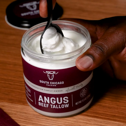 A hand holding a spoonful of white beef tallow from a clear jar with a dark burgundy and white label that reads 'South Chicago Packing Angus Beef Tallow.' The jar sits on a wooden surface, with the lid placed nearby.