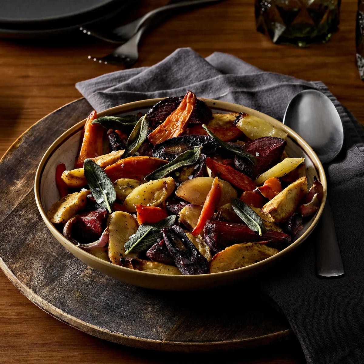 A bowl of roasted root vegetables and beets with sage on a wooden board, set on a dark tablecloth with a spoon and napkin.