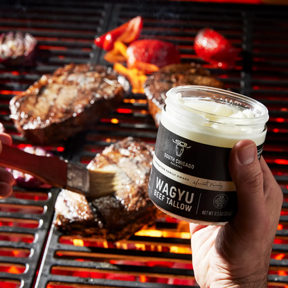 A hand holds a clear jar of South Chicago Creamery Wagyu Beef Tallow basting spray over a grill, with a brush applying the white tallow to a steak. Bright red strawberries and glowing orange grill grates are visible in the background.