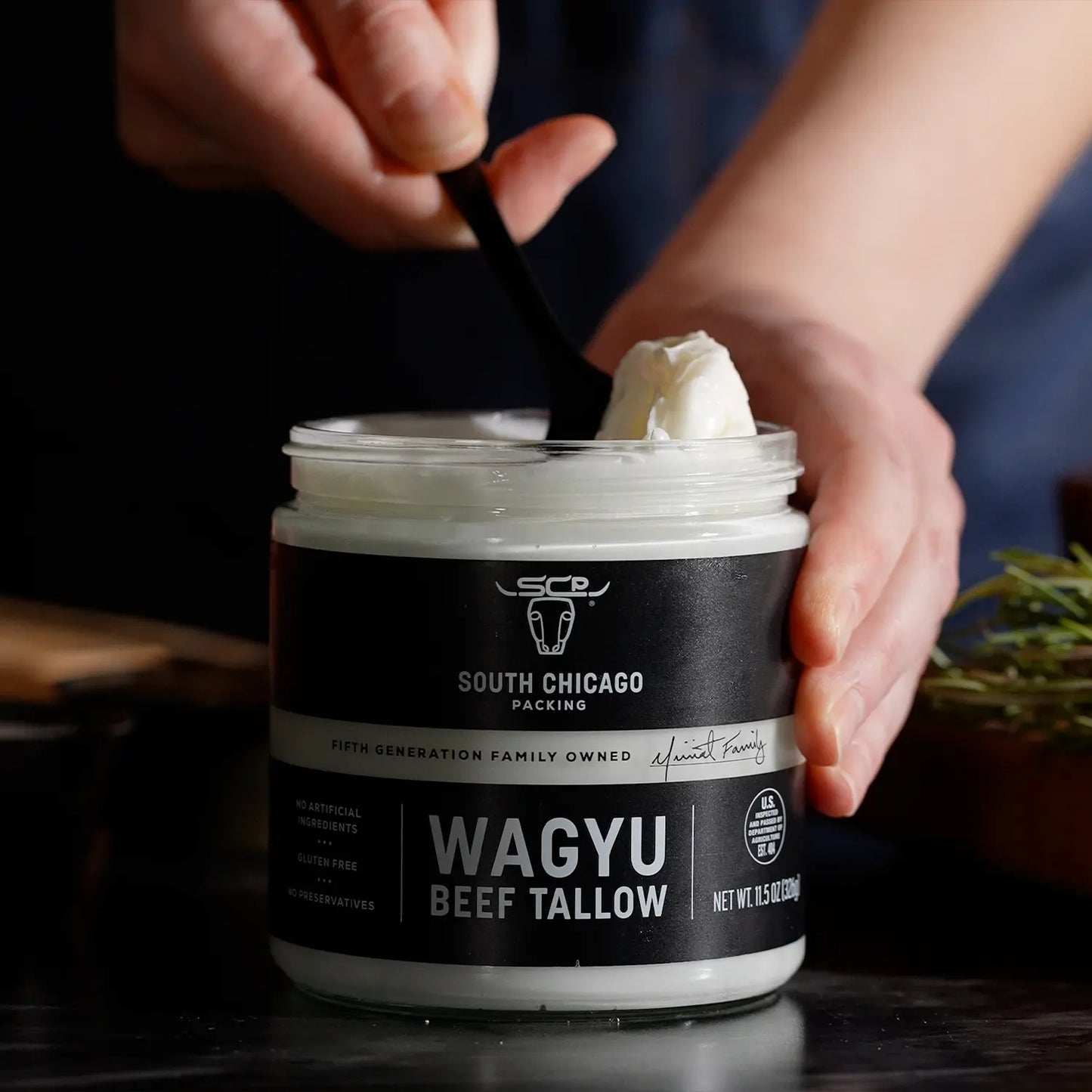 A person's hands holding a jar of South Chicago Creamery Wagyu Beef Tallow ghee, with a black spoon scooping some of the white ghee from the top. The jar has a black label with white text, set against a dark, moody background with a hint of greenery on the side.