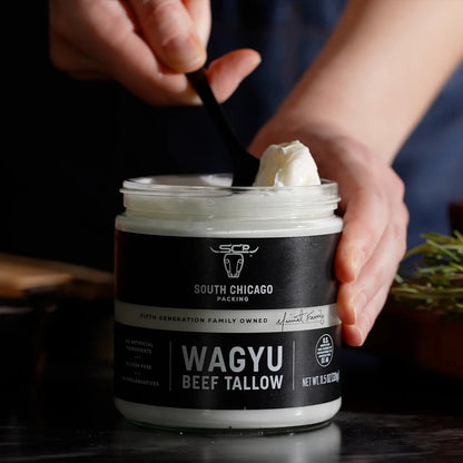 A person's hands holding a jar of South Chicago Creamery Wagyu Beef Tallow ghee, with a black spoon scooping some of the white ghee from the top. The jar has a black label with white text, set against a dark, moody background with a hint of greenery on the side.