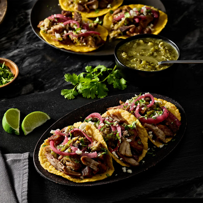 Three soft corn tacos topped with shredded meat, pickled red onions, cilantro, and crumbled cotija cheese, served on a dark oval platter with lime wedges and a bowl of green salsa on a dark tabletop.